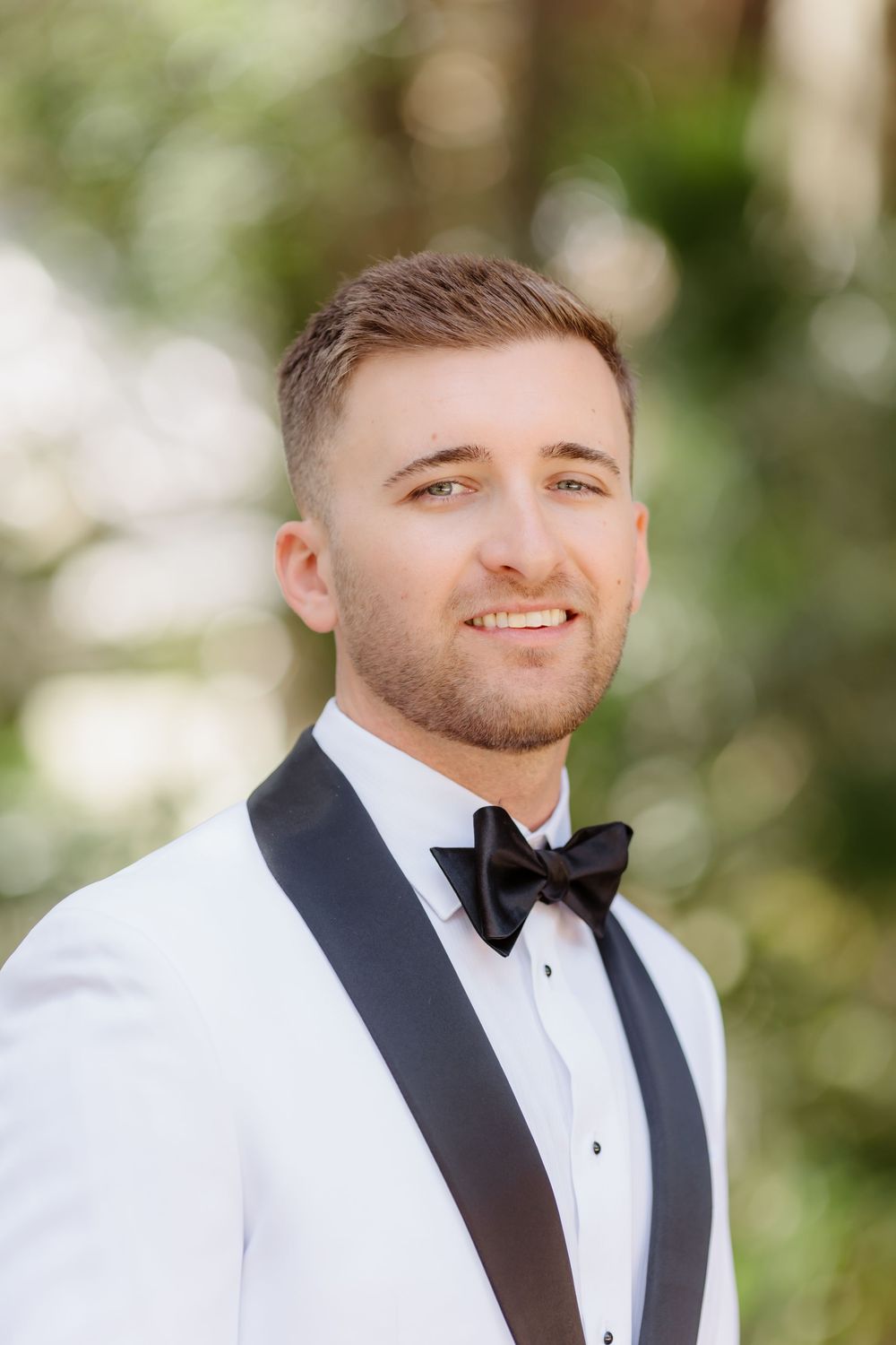 Handsome groom smiles in white tuxedo jacket with black bow tie and lapels at outdoor wedding.