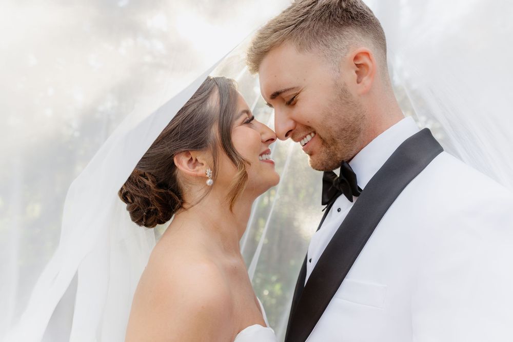 Bride and groom share a romantic moment under a flowing veil, smiling at each other on their wedding day.