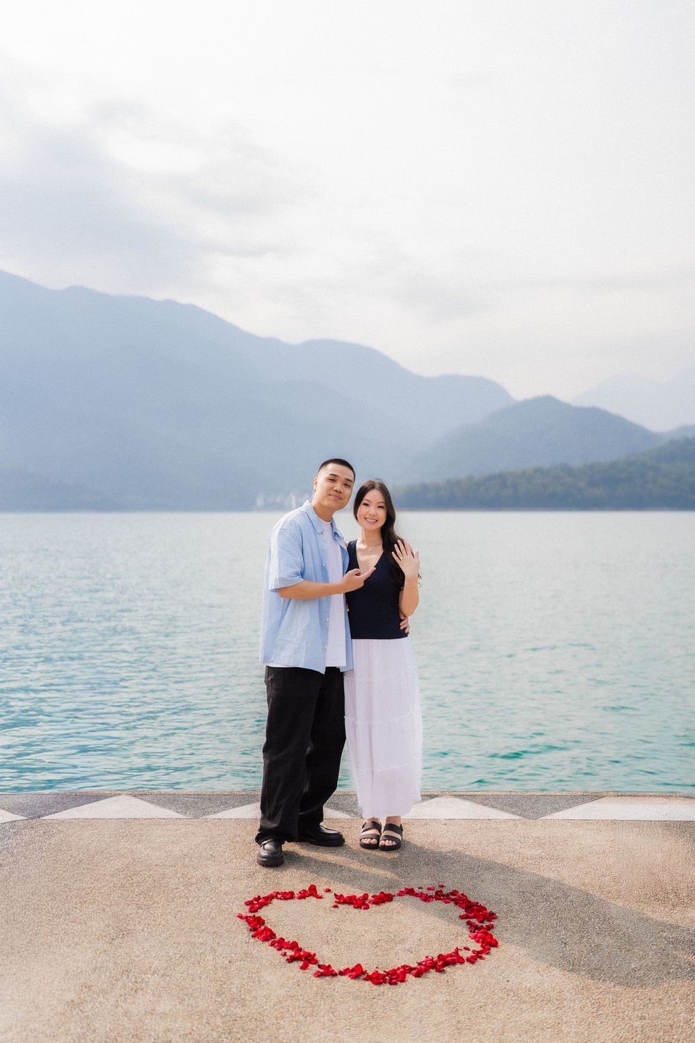 Couple posing by a lakeside heart made of red rose petals with mountains in the background.