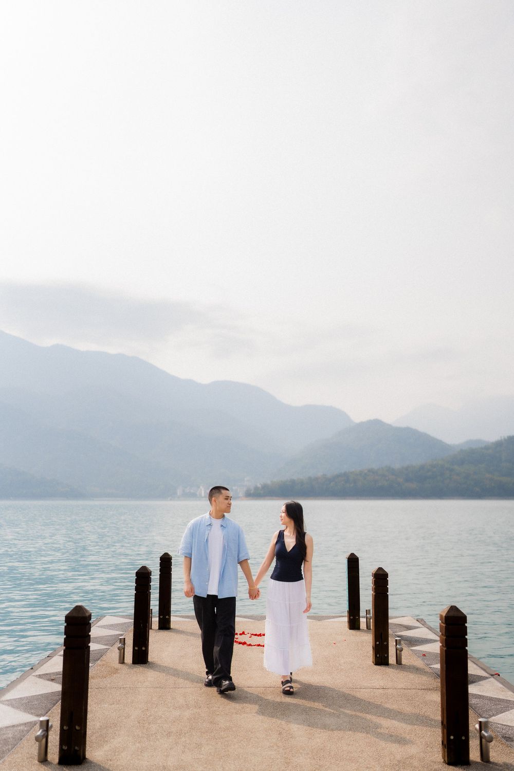 Couple holding hands on a lakeside pier with misty mountains and turquoise water in the background.