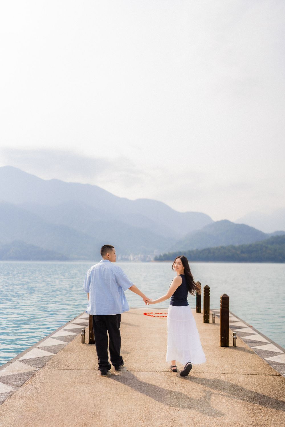 Couple holding hands on a scenic lakeside pier with misty mountains in the background.