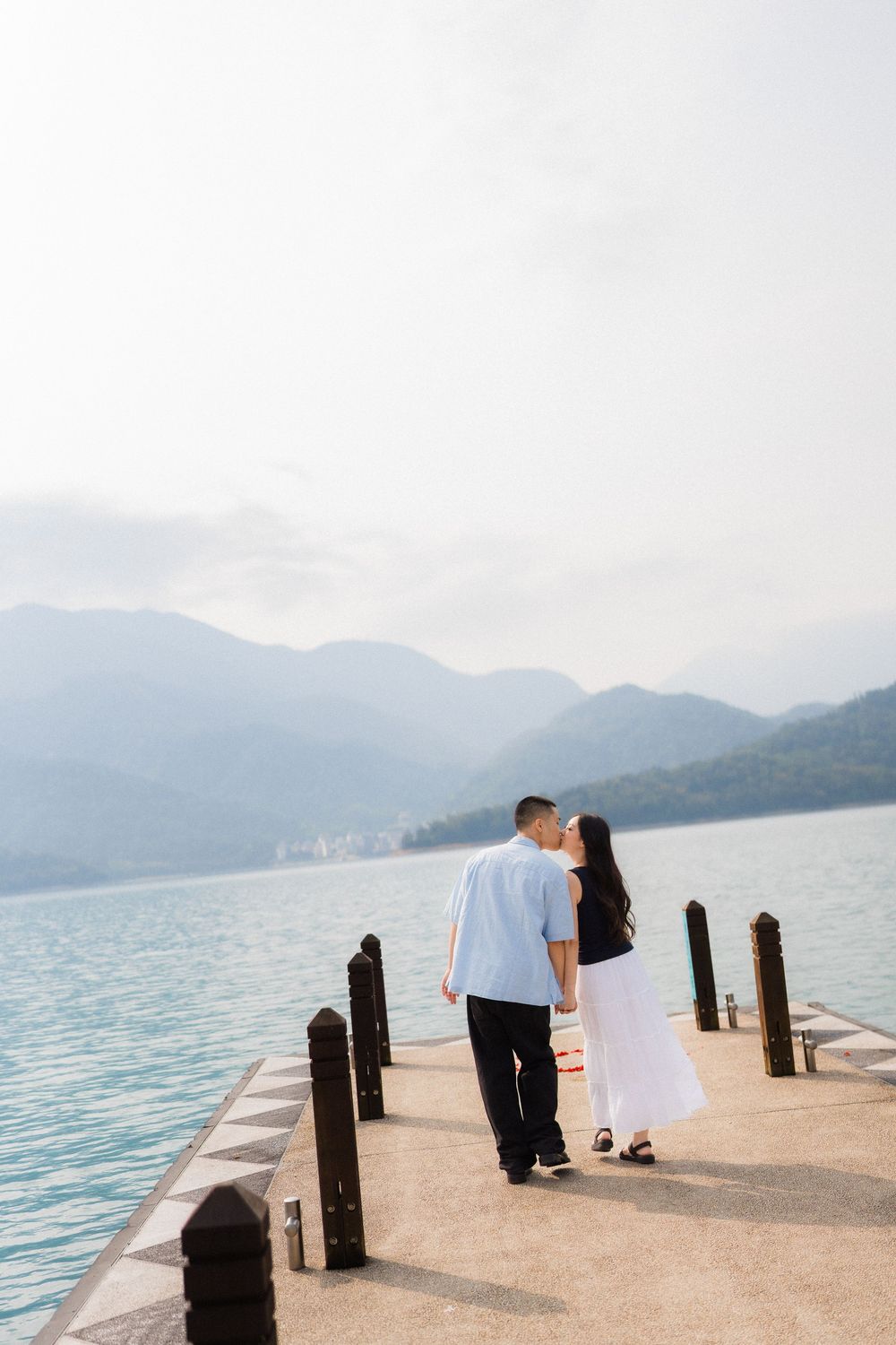 Couple sharing a romantic kiss on a lakeside pier with misty mountains and calm blue waters in the background.