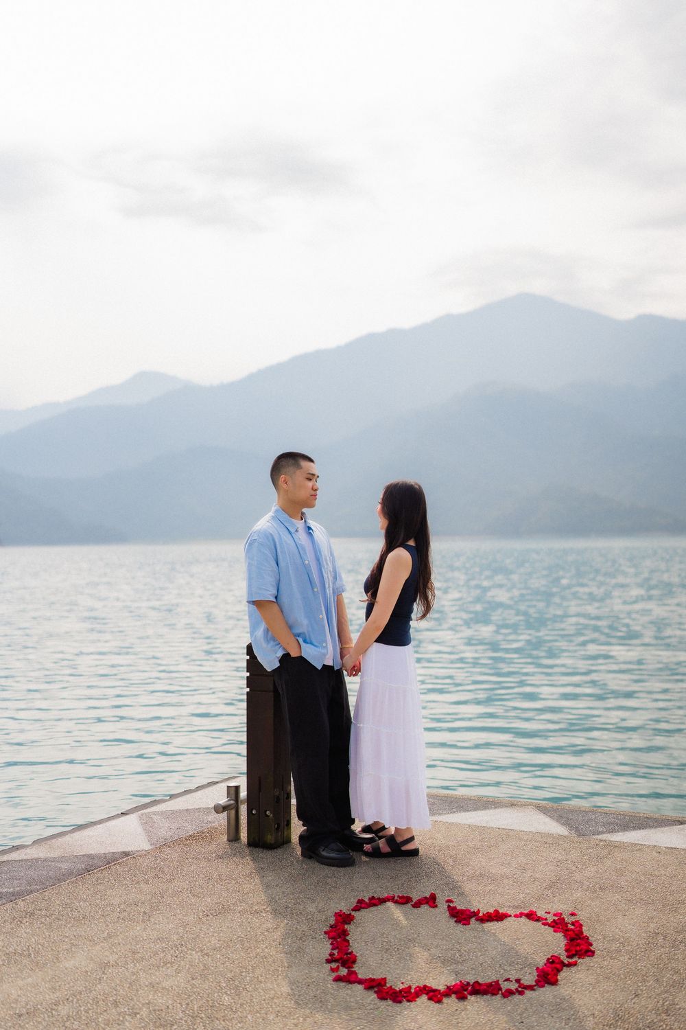 Couple holding hands on a lakeside dock with a rose petal heart on the ground and misty mountains behind.