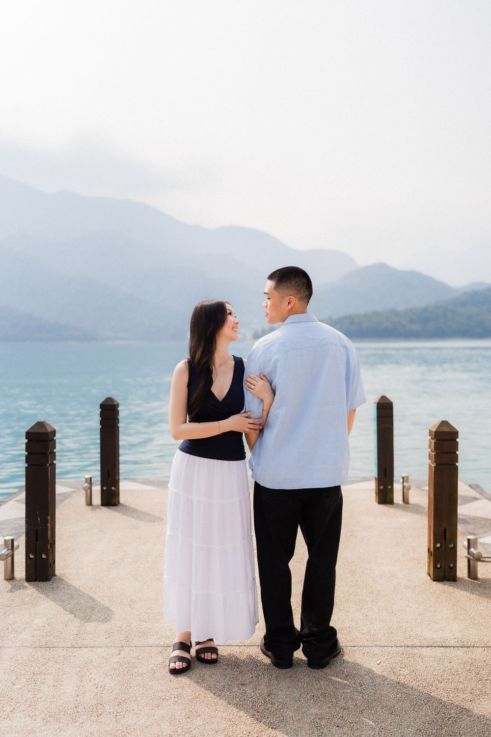 Couple embracing on a lakeside pier with turquoise water and misty mountains in the background.