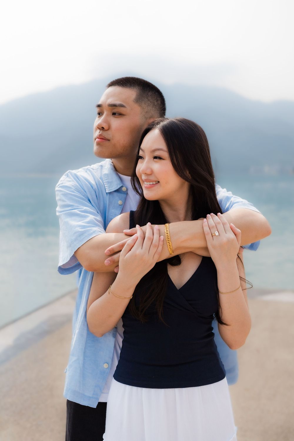 A young Asian couple embracing near a waterfront with mountains in the background, woman smiling warmly.