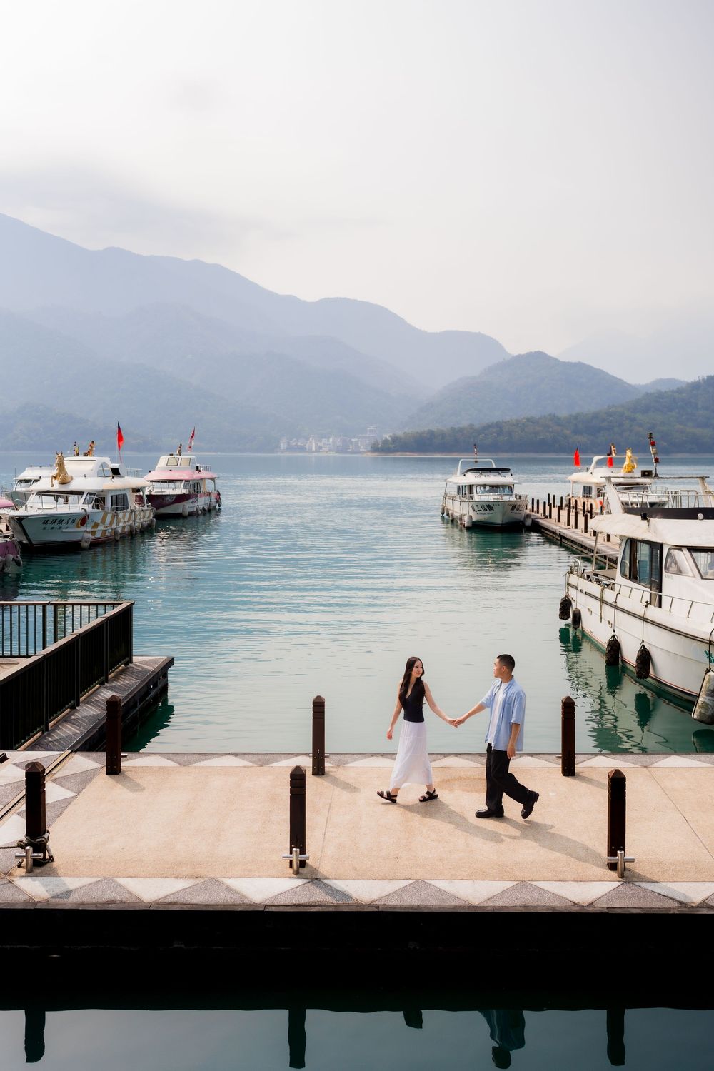Couple holding hands on a marina dock with boats and scenic mountain lake views.