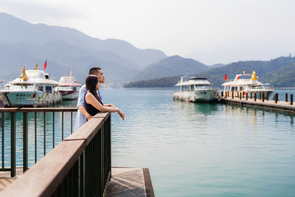 Two people stand on a wooden dock overlooking a serene lake with moored boats and misty mountain backdrop.