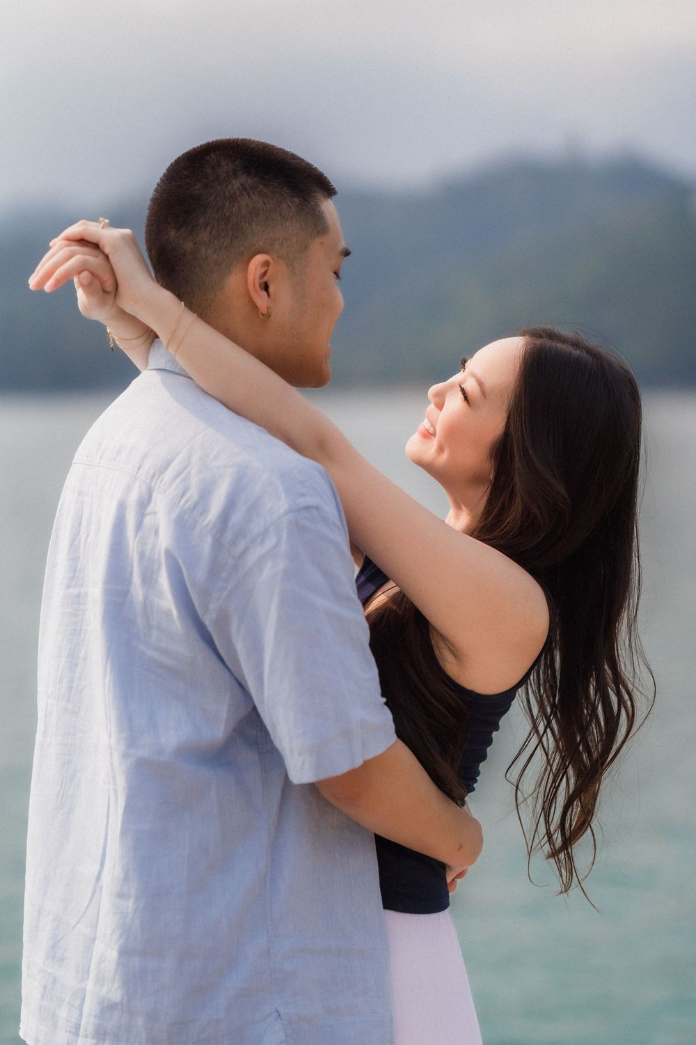 Romantic couple embracing by a serene lake, woman smiling with arms around mans neck outdoors.