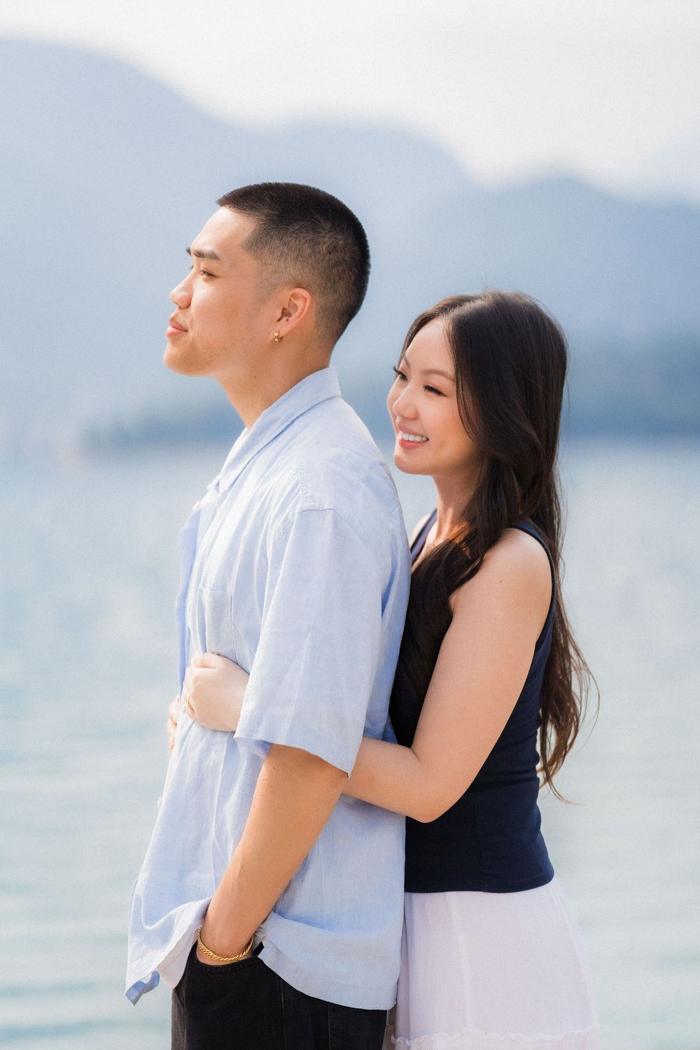 A smiling Asian couple embraces by a serene lake with mountains, woman in navy top hugs man in light blue shirt.