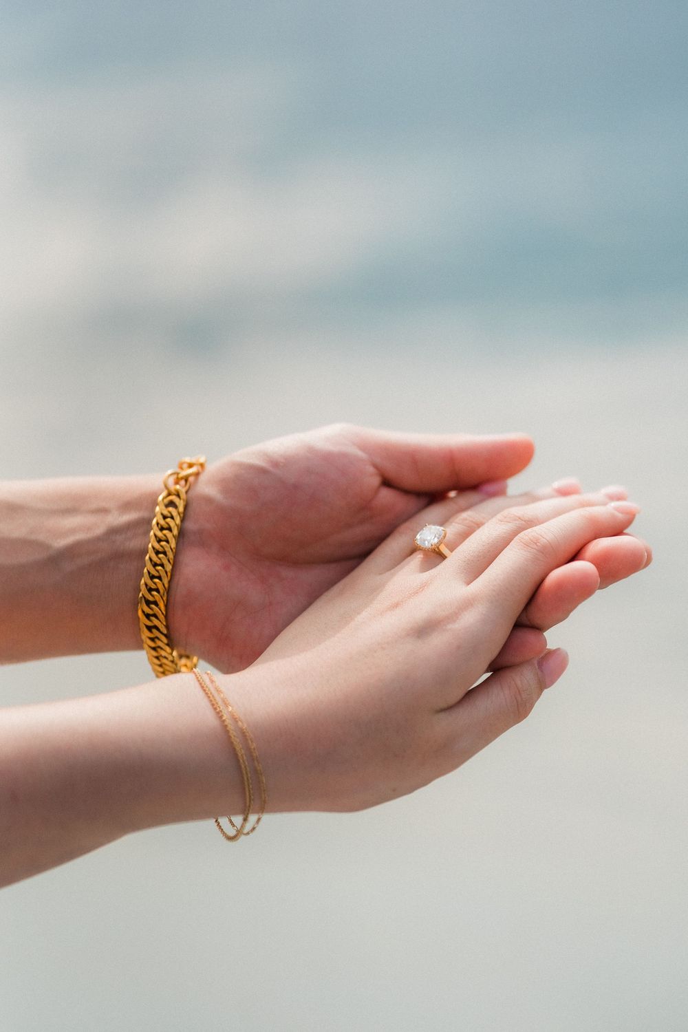 Two hands cupped together near the ocean, adorned with gold chain bracelet and delicate ring, holding a small crystal.