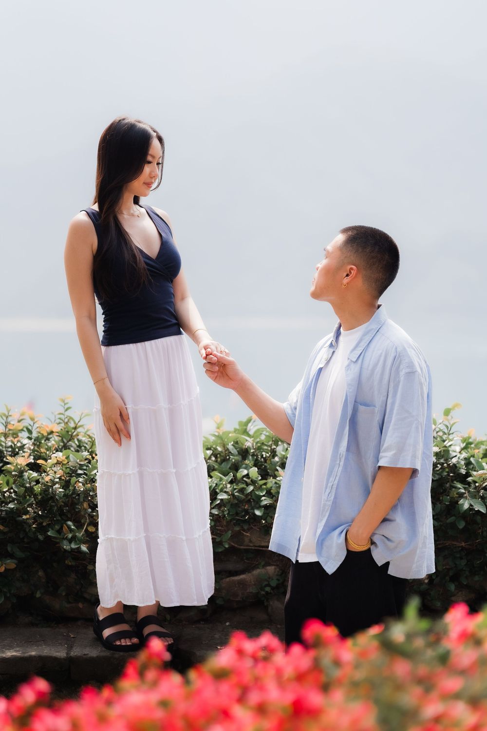 Man on one knee holding womans hand in a romantic proposal scene with flowers and lake views.