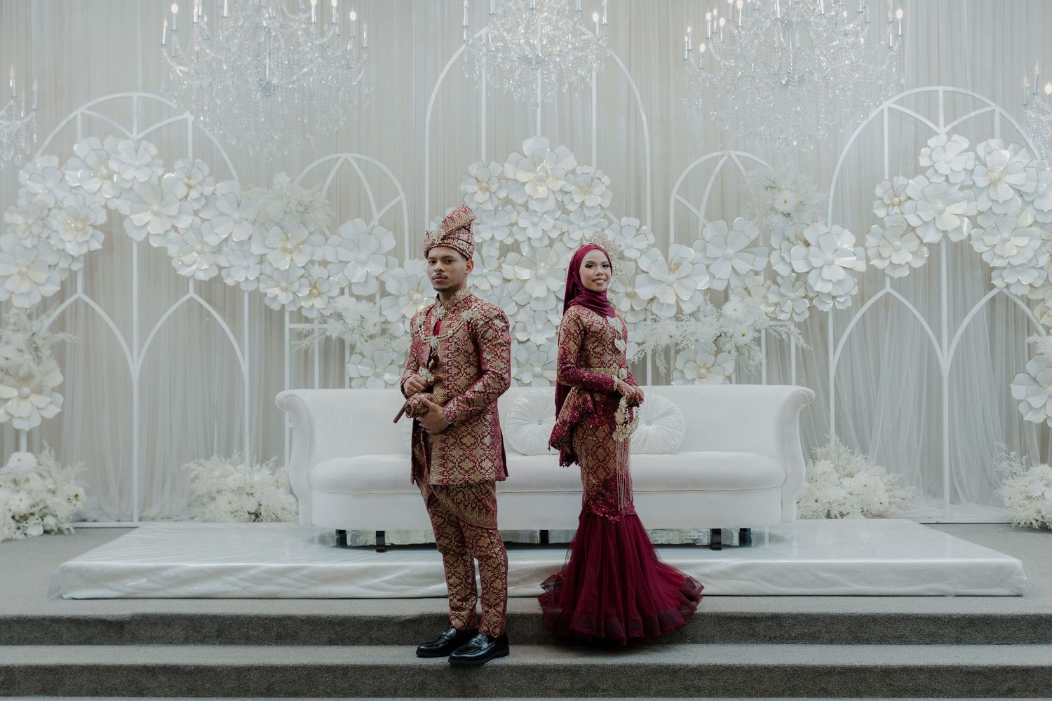 Indonesian wedding couple in traditional red and gold attire posing before an elegant white floral backdrop at a formal ceremony.