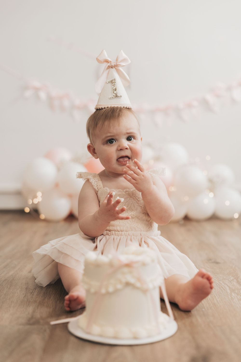 Baby girl in tutu and birthday hat smashes first birthday cake surrounded by white balloons.