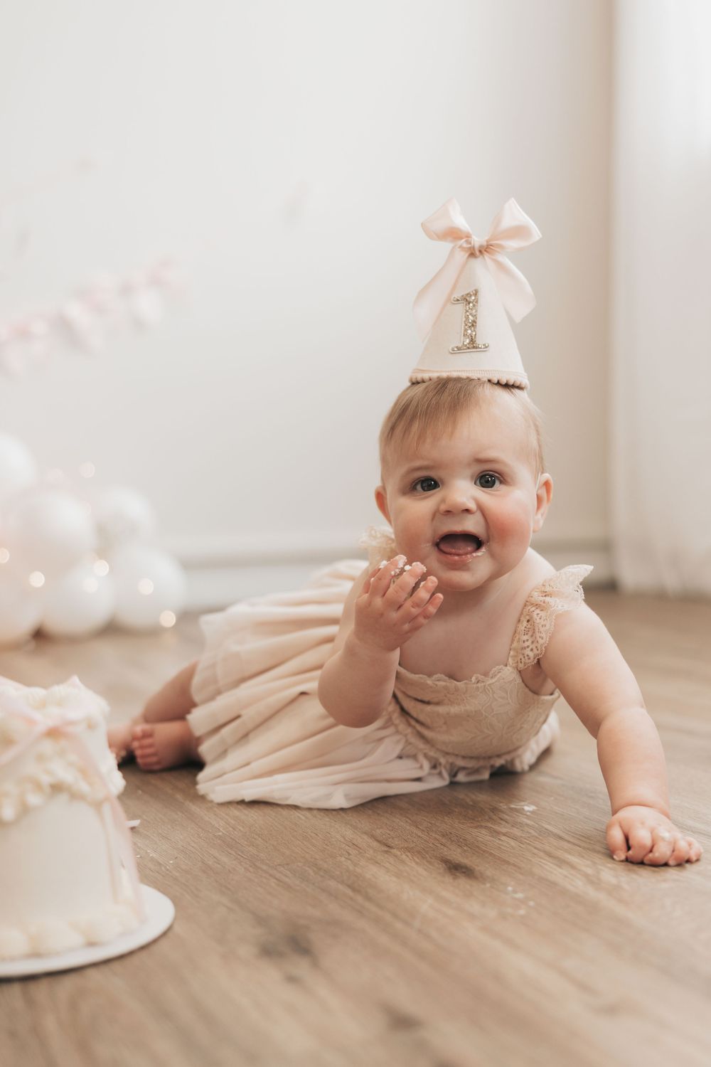 Adorable baby girl in blush romper and number one party hat crawling toward white smash cake on wood floor.