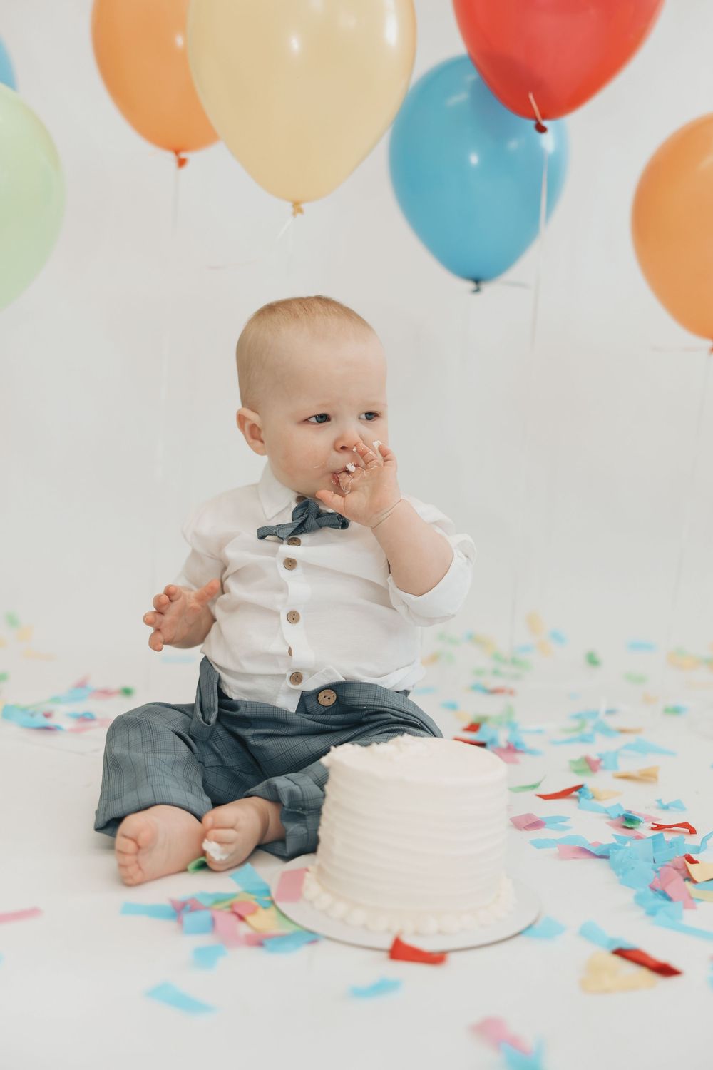 Baby boy in white shirt enjoys first birthday cake smash surrounded by colorful balloons and confetti.