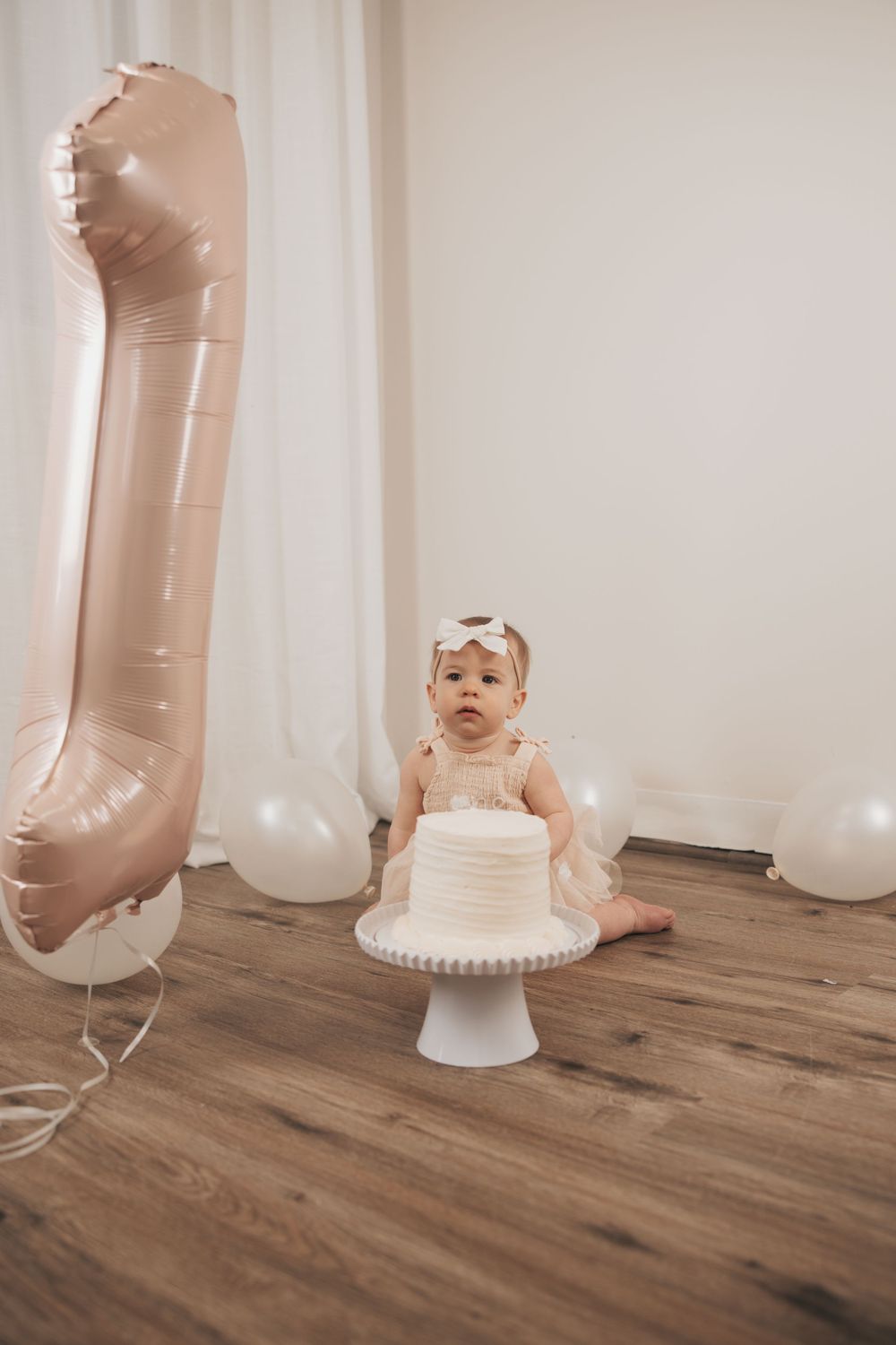 Baby girl in white dress sits before smash cake at first birthday party with rose gold number one balloon.