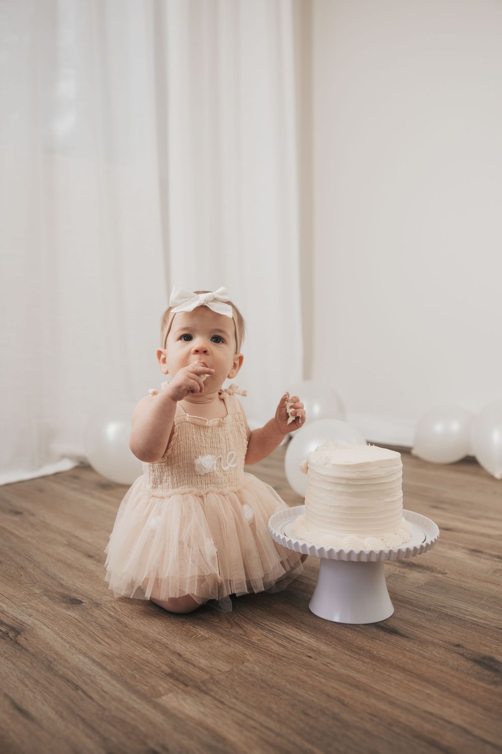 Baby girl in pink tutu dress with bow headband tastes her first birthday smash cake at white decorated party.