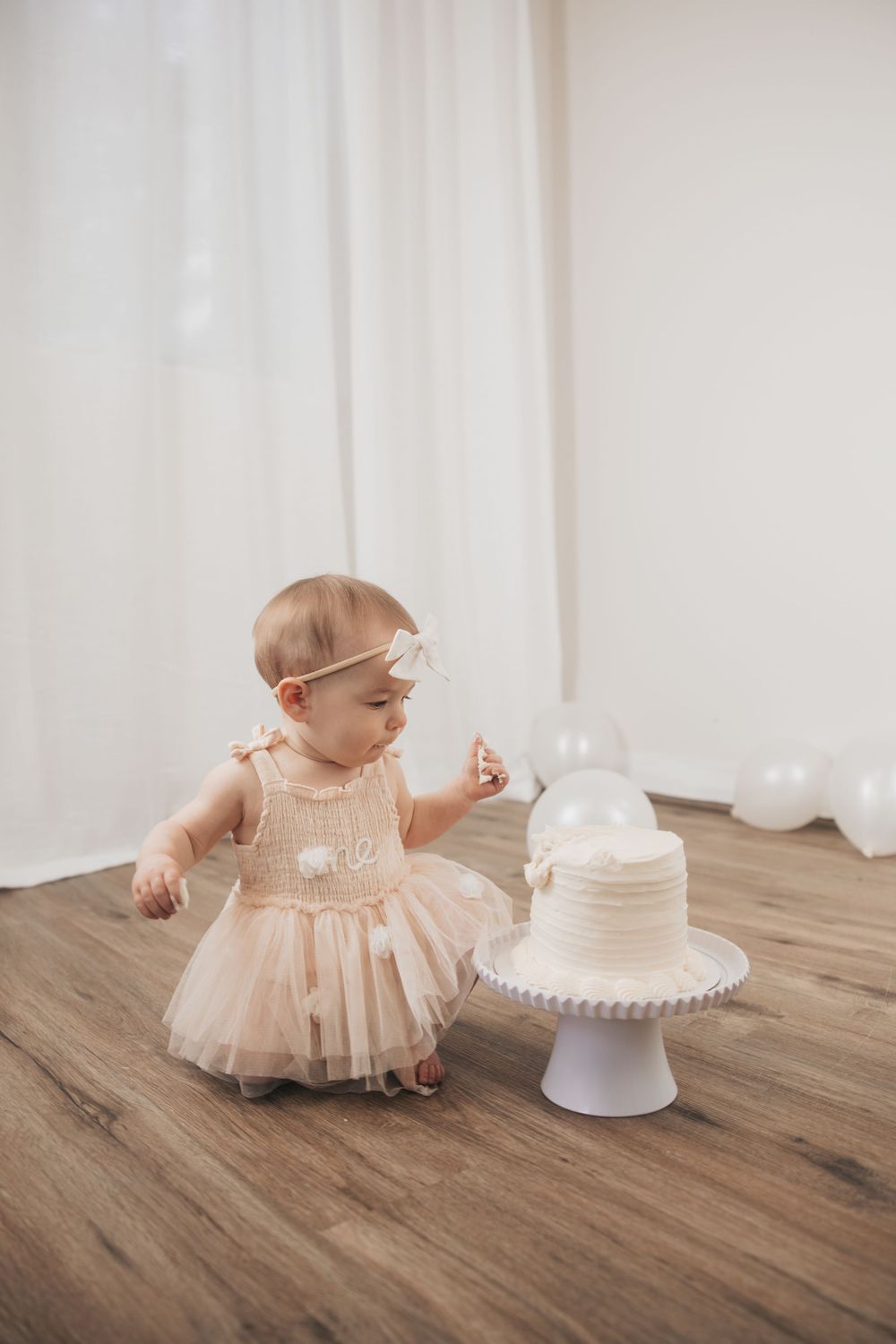 Baby girl in pink tutu dress reaching toward white first birthday cake on stand with balloons in bright white room.