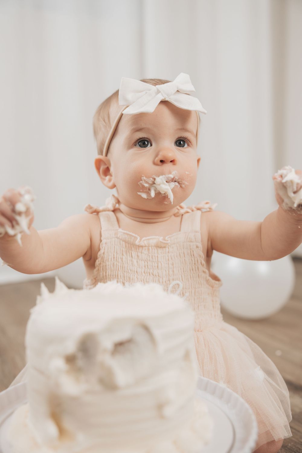 Baby girl in pink dress with bow headband smashing her first birthday cake, covered in white frosting.