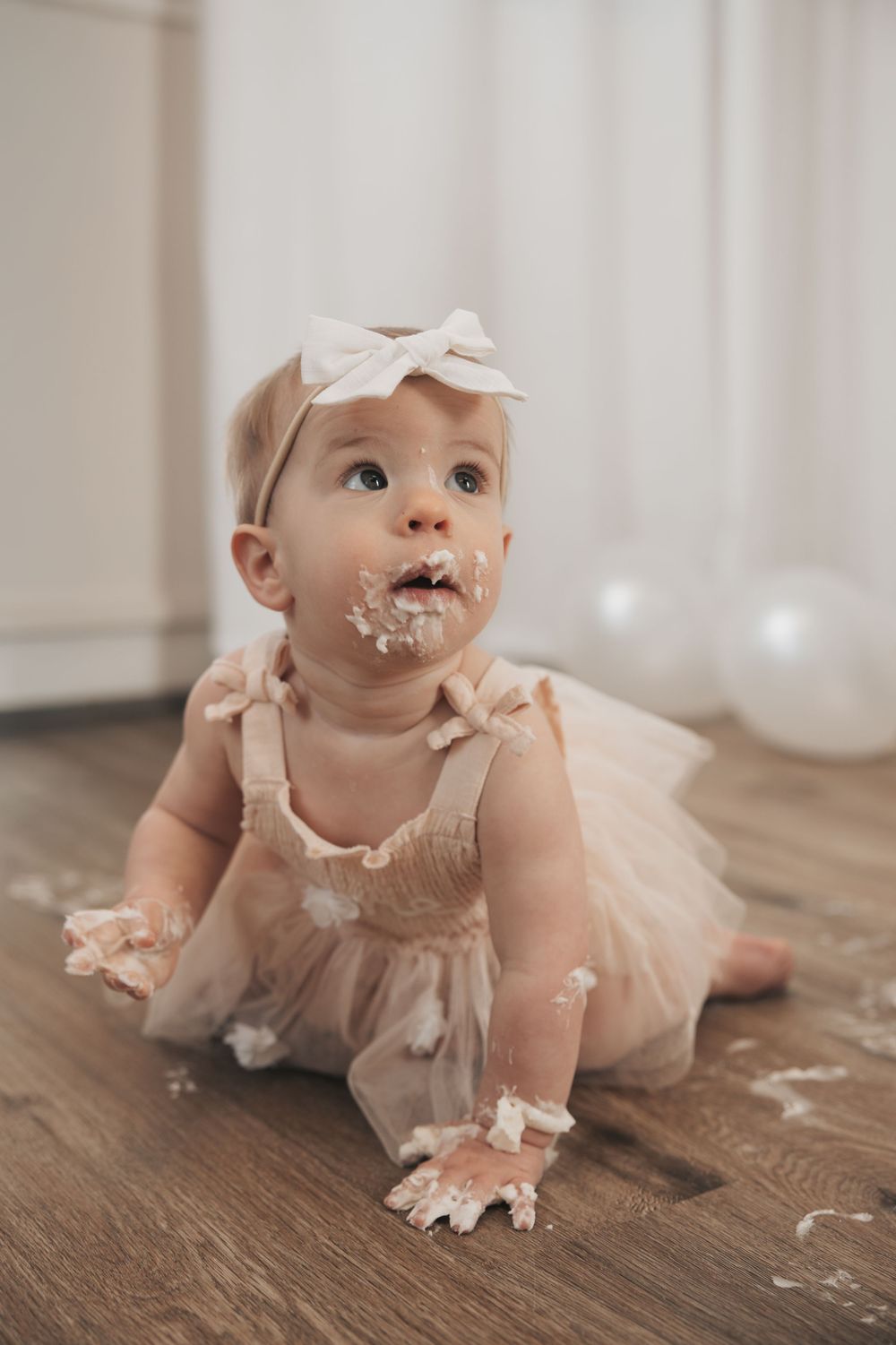 Baby girl in pink tutu dress with bow headband sitting on floor with cake smash frosting on face and hands.