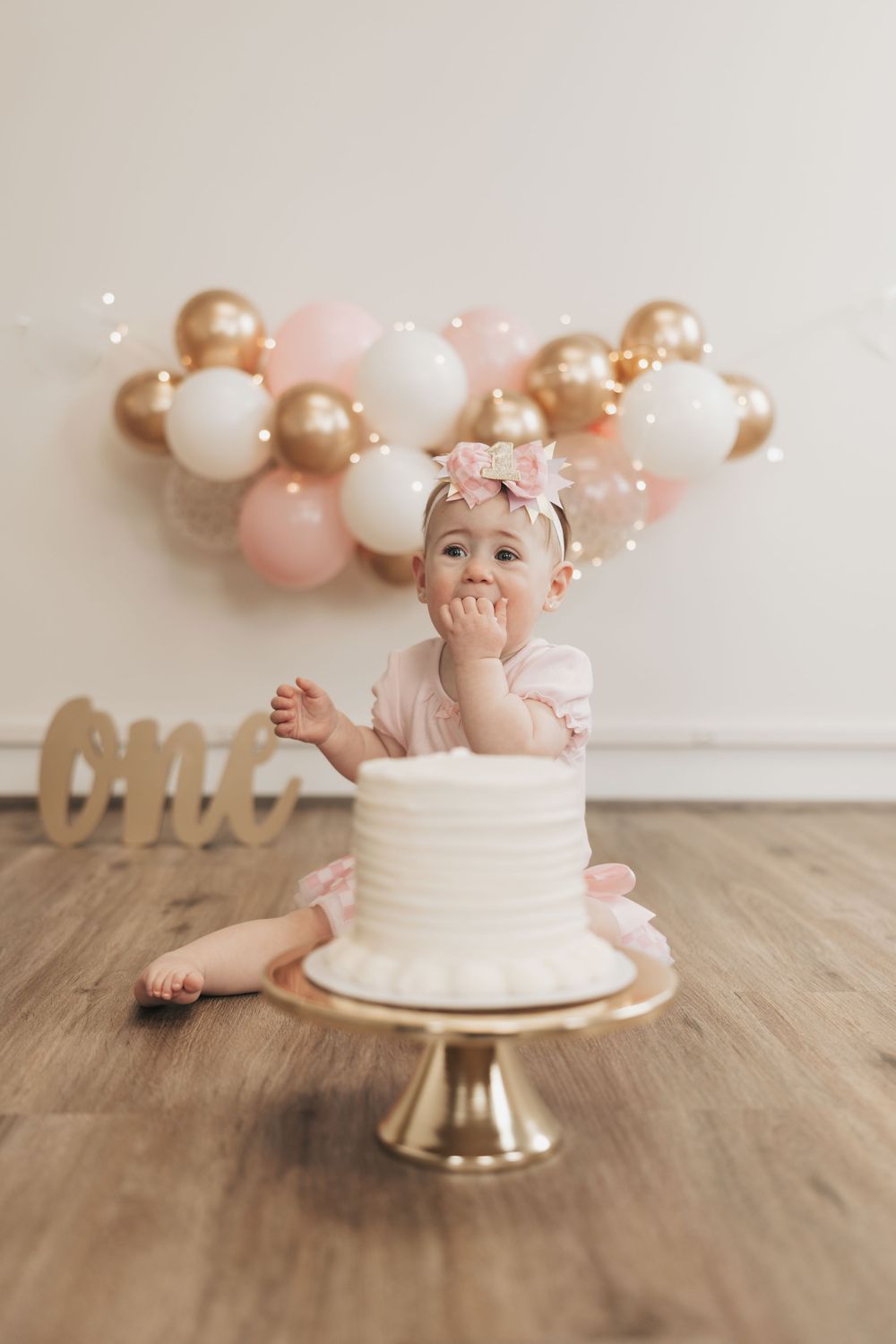 Baby girl in pink tutu sits before white cake on gold stand, celebrating first birthday with balloons backdrop.