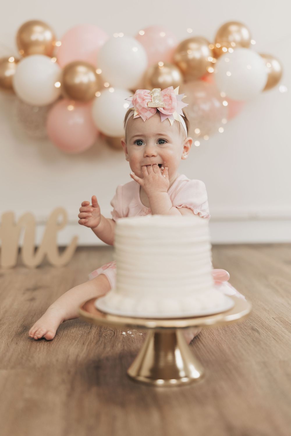 Baby girl in pink bow headband smiles at her first birthday cake smash with gold and pink balloon backdrop.