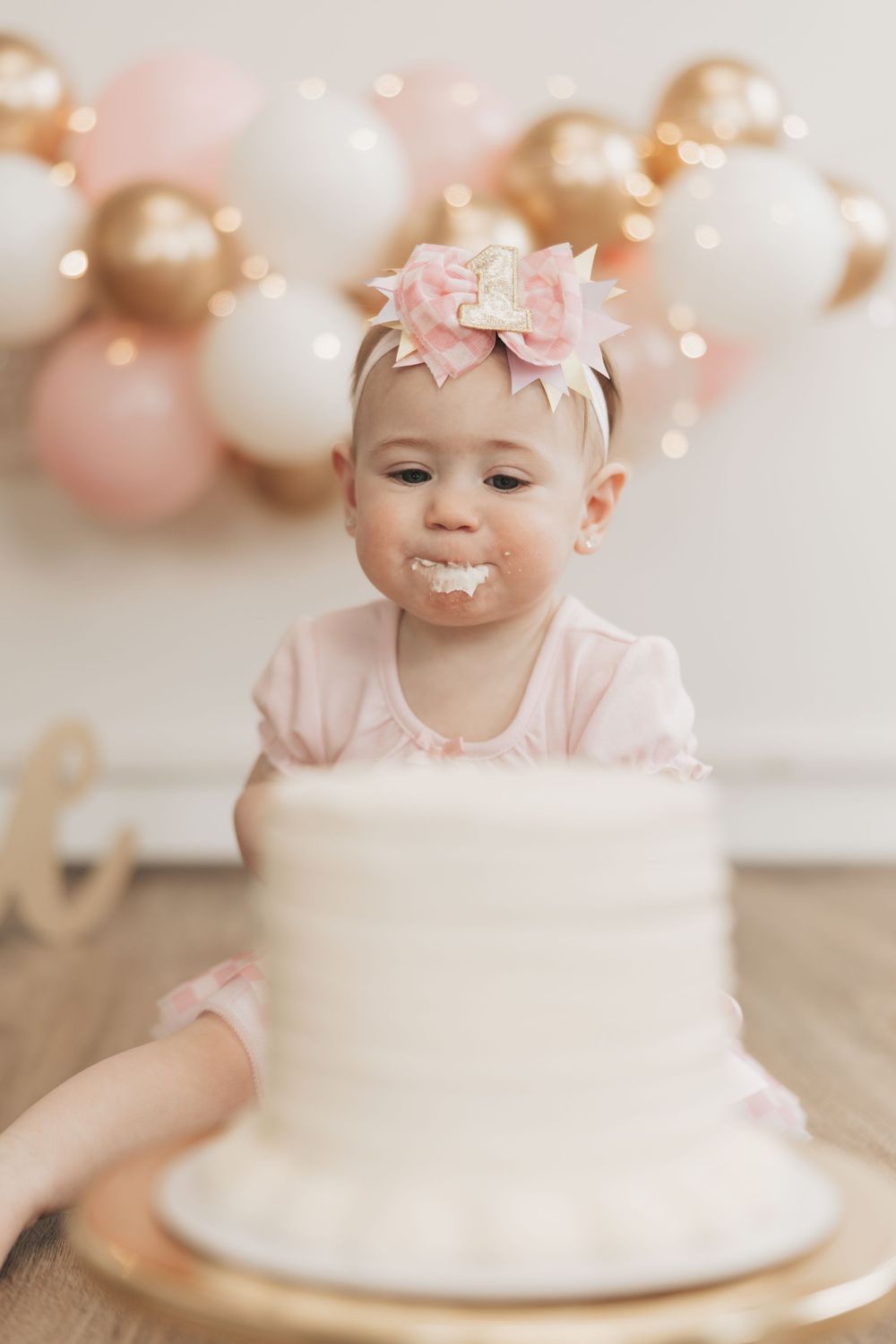 Baby girl in pink outfit celebrates first birthday with white cake, wearing a number one crown bow headband.