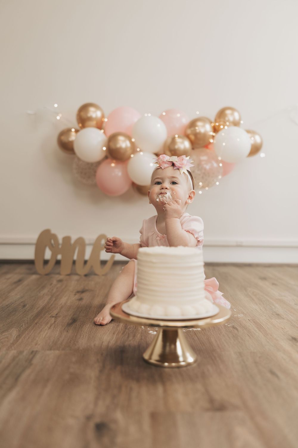 Baby girl in pink dress celebrates first birthday with white cake, gold stand, and pink and gold balloon garland backdrop.