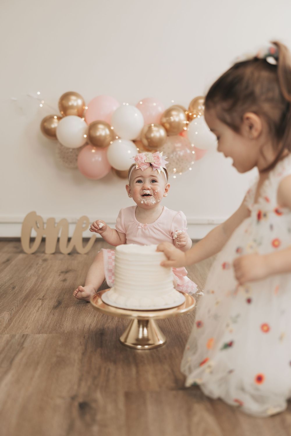 Baby girl celebrates first birthday with smash cake, older sister nearby, pink balloons and “one” sign in background.