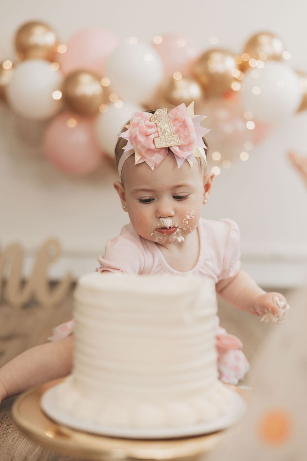 Baby girl in pink bow headband smashes white birthday cake at first birthday party with gold and pink balloon backdrop.