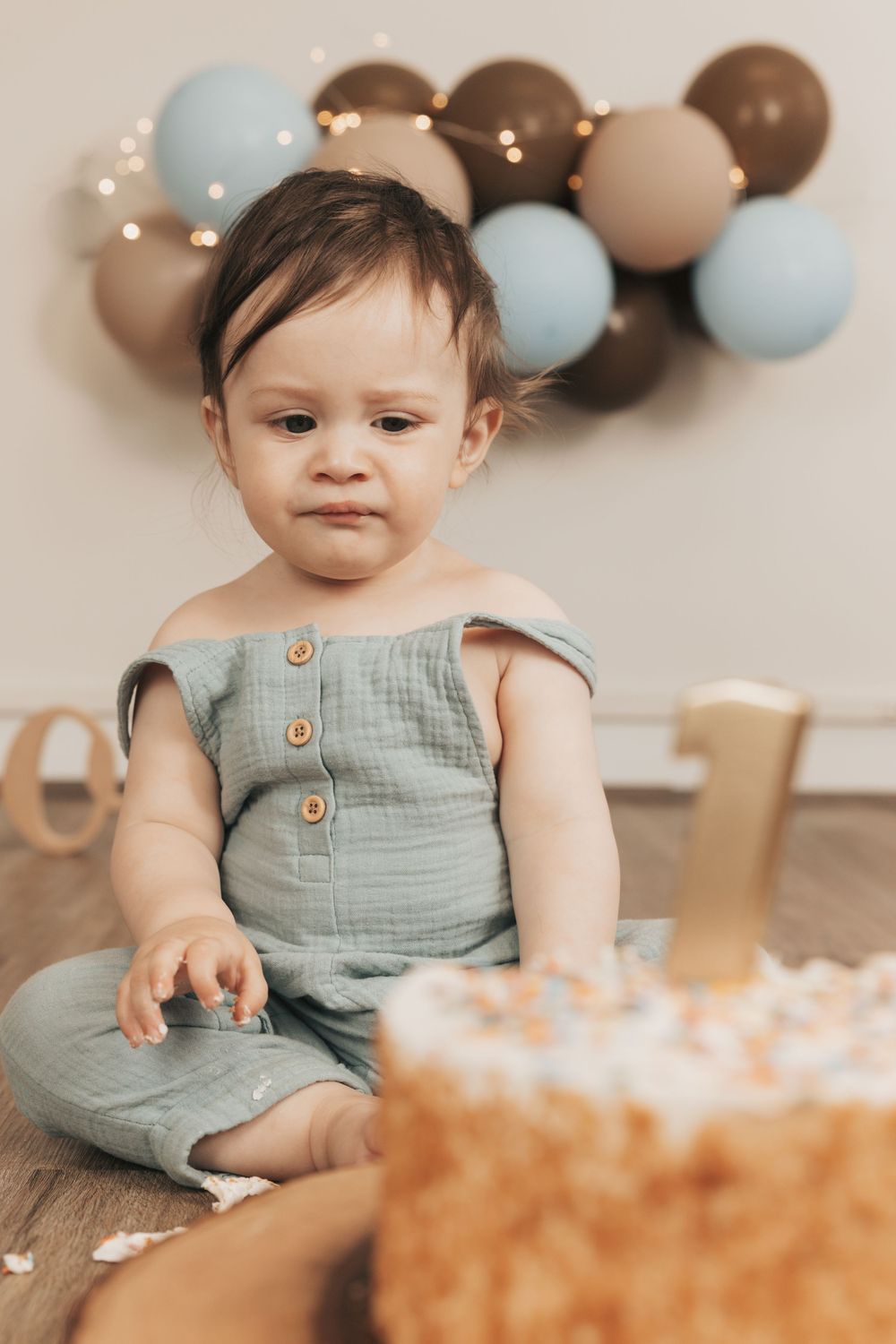 Baby in teal romper sits beside a first birthday cake, with blue and brown balloons on wall behind.