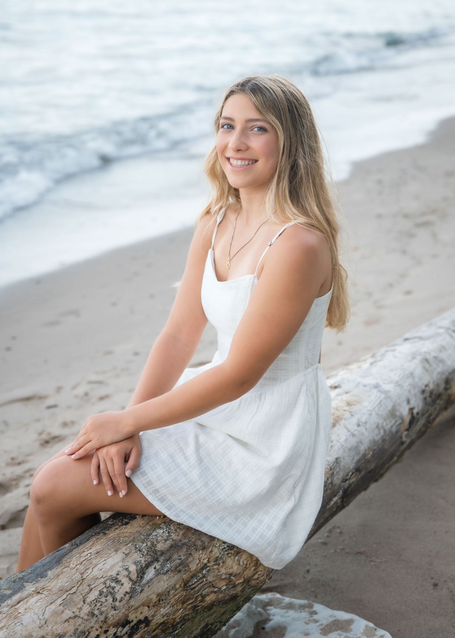Young blonde woman in white top and blue jeans sits barefoot on rustic wooden steps surrounded by lush greenery.