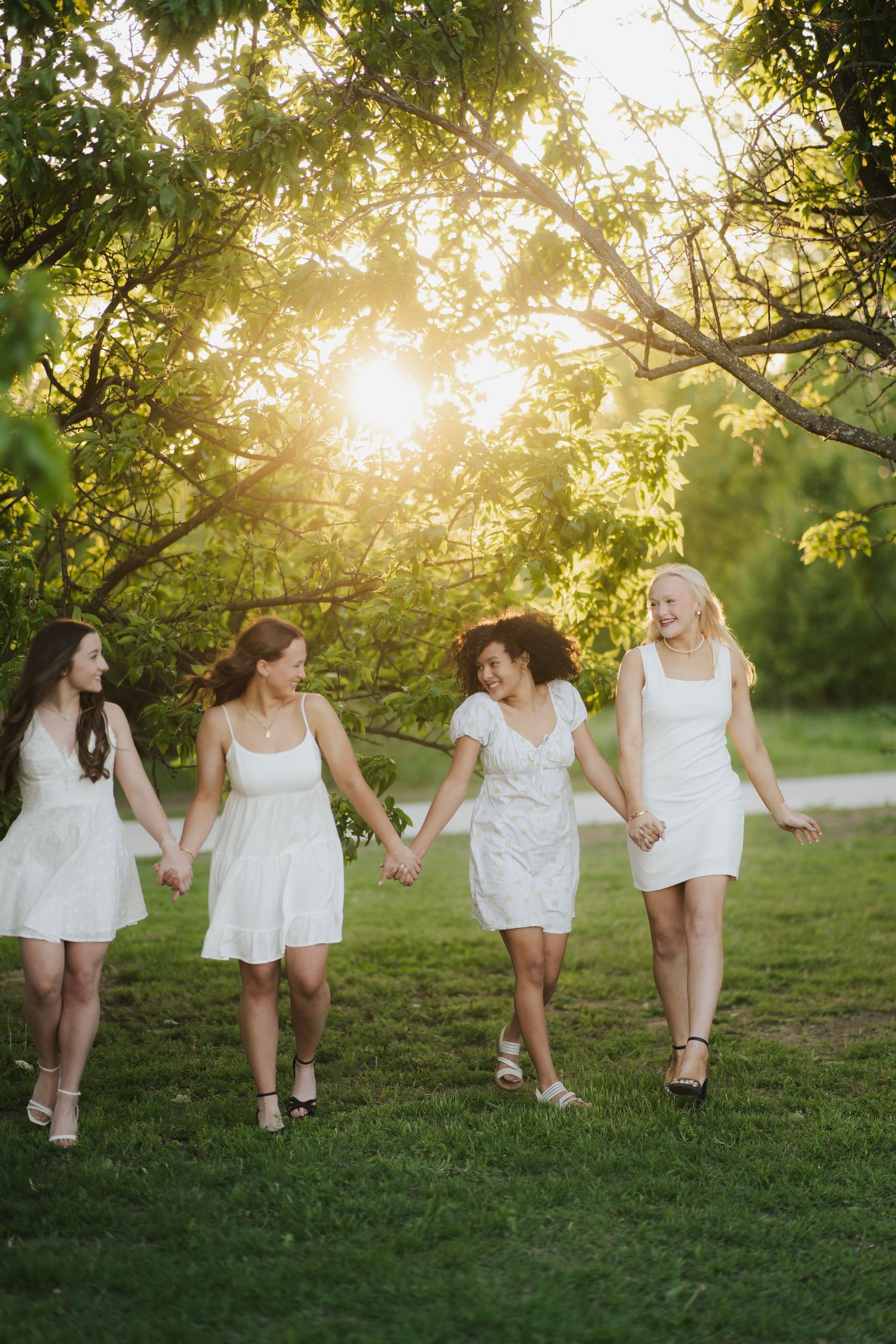 Four young women in white dresses hold hands walking together through a sunlit green park.