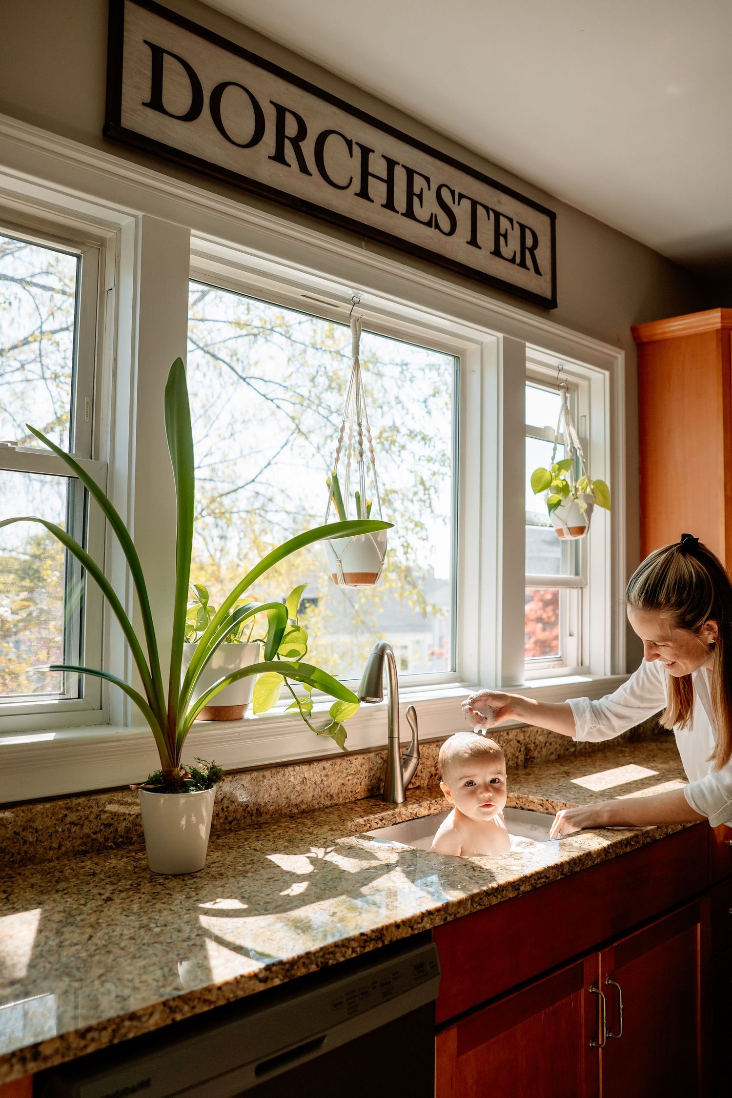 Mother bathing smiling baby in kitchen sink near windows with plants and Dorchester sign above.