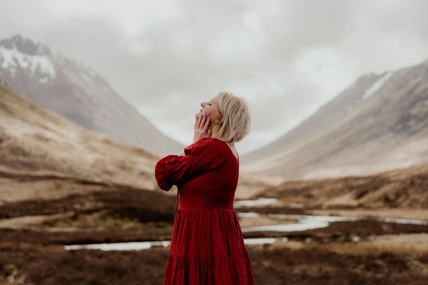 Woman in red dress gazing upward in Scottish Highland valley with misty mountains backdrop.
