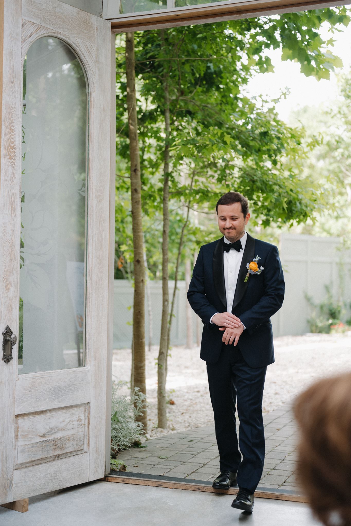 A groom in a black tuxedo with boutonniere smiles while standing in an elegant doorway surrounded by lush greenery.