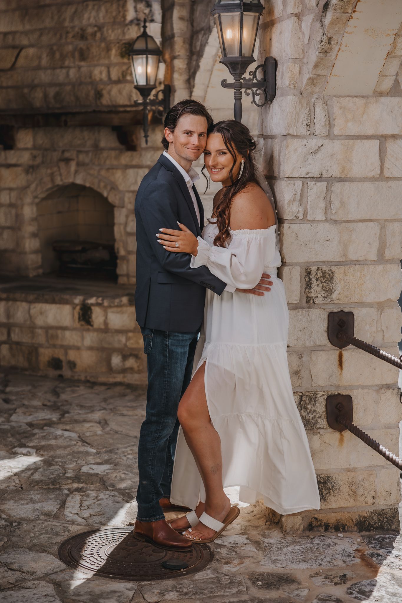 Couple embracing outdoors near stone fireplace, woman in white off-shoulder dress, man in navy blazer and jeans.