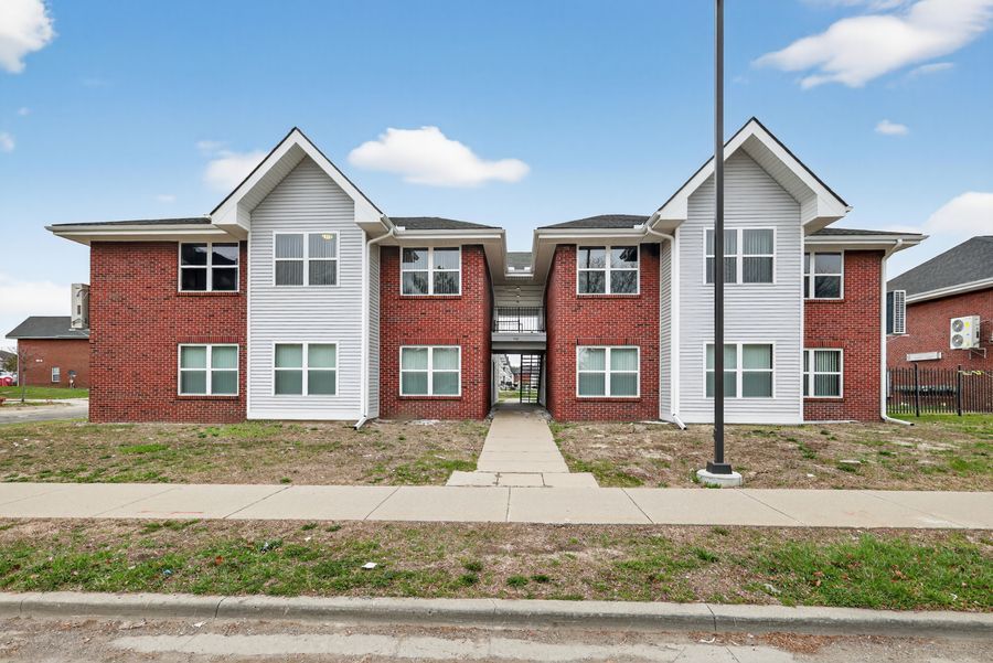 Modern two-story brick apartment building with white trim, manicured lawn, and central entrance walkway under a partly cloudy sky.