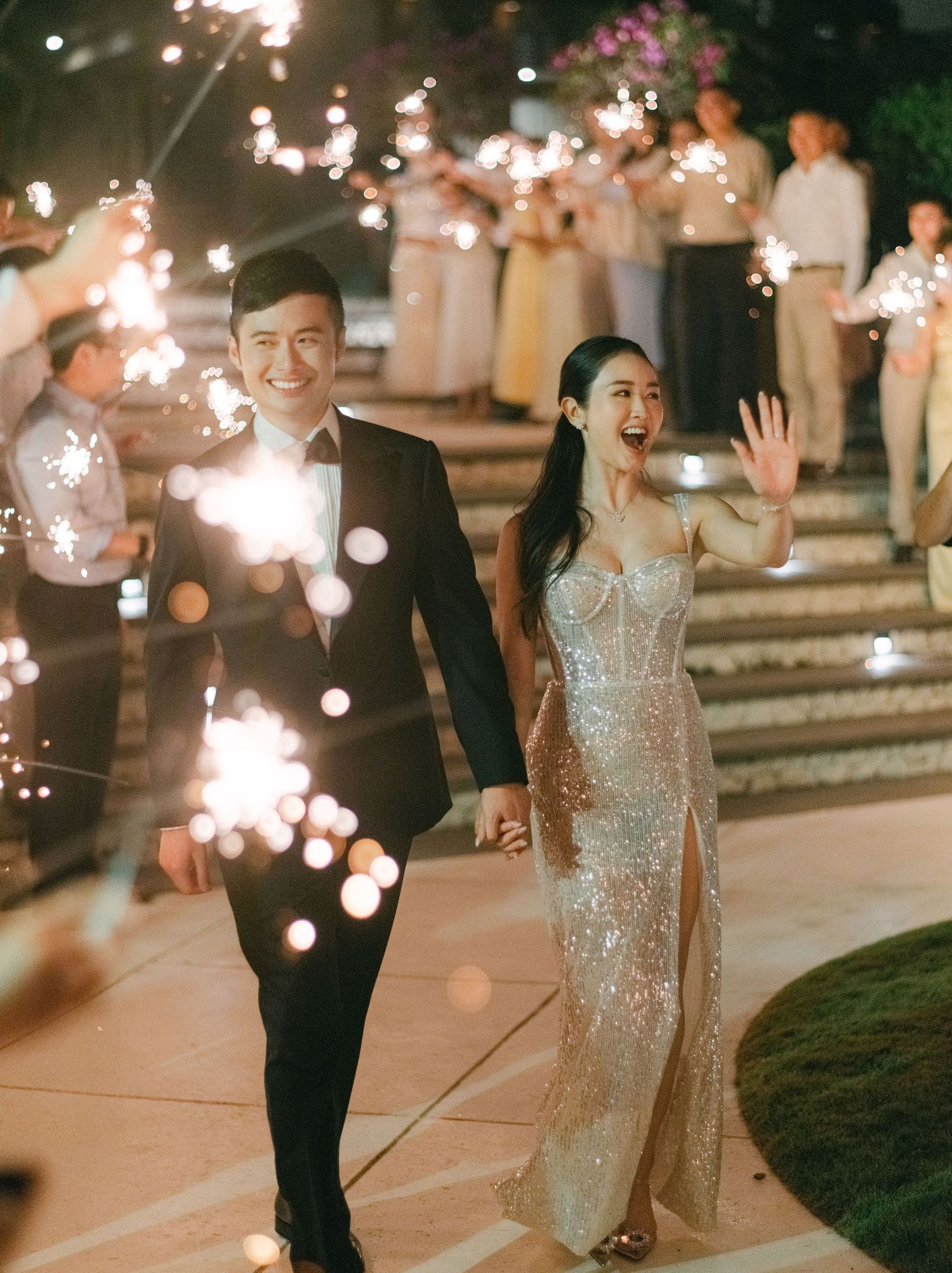Newlywed couple walking through sparkler exit at night wedding reception, bride in sequin gown, groom in black tuxedo.