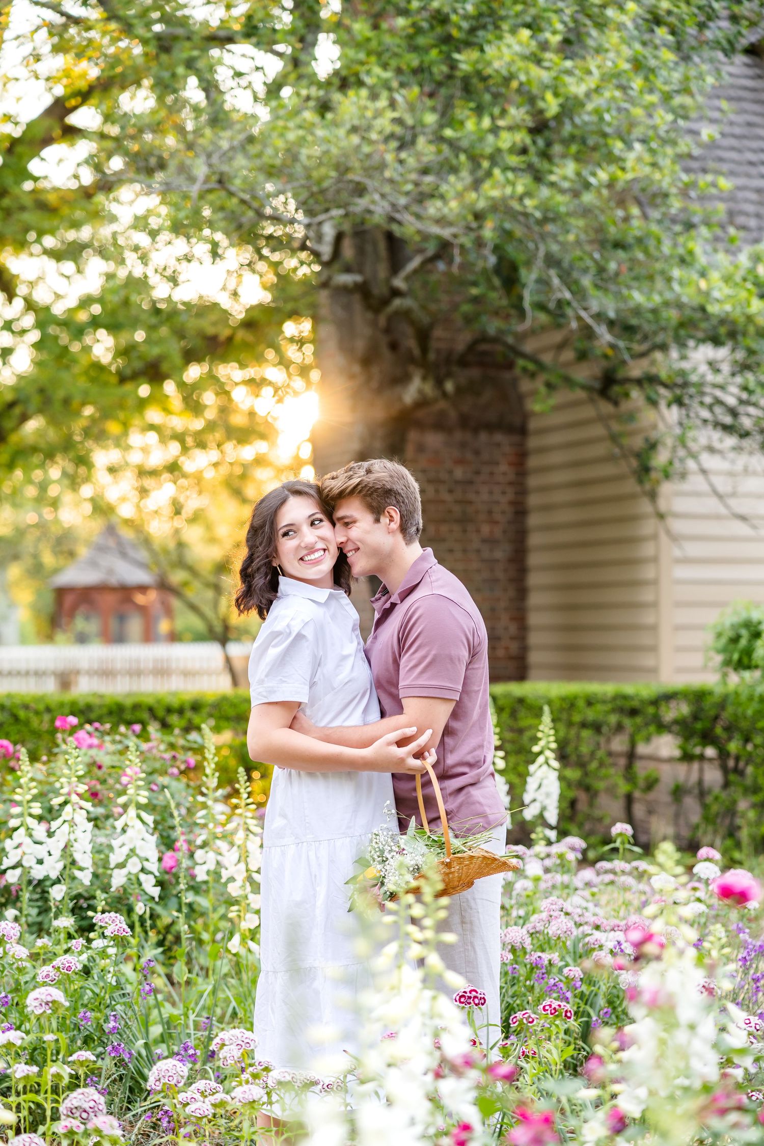 Photography of a newly engaged couple snuggling in a flower garden in Colonial Williamsburg illuminated by golden hour glowing sunlight.