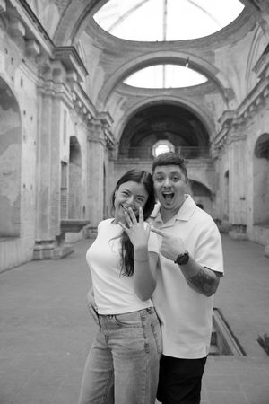 Black and white photo of a happy couple showing off an engagement ring inside a historic arched stone building.