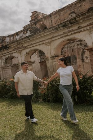 Couple holding hands and walking on green grass in front of ancient stone ruins with arched windows in Antigua, Guatemala.