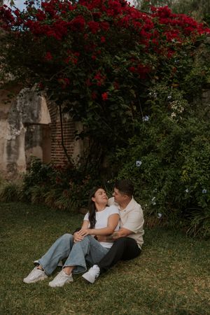 Romantic couple sitting on grass beneath vibrant red bougainvillea flowers at historic ruins.