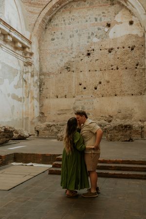 Couple sharing a romantic kiss in a historic stone ruins courtyard, woman in green dress, man in khaki shorts.