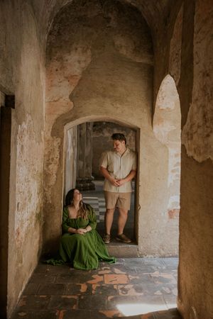 Couple poses in ancient stone archway, woman in green dress sits on floor looking up at man in doorway.