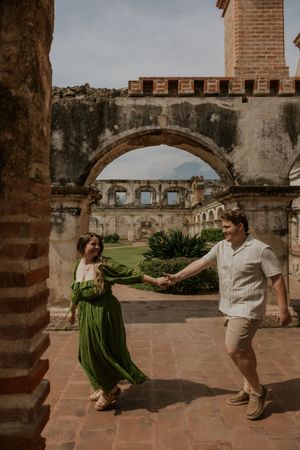 Couple holding hands and dancing among ancient stone arches and ruins, woman in flowing green dress, outdoor courtyard.