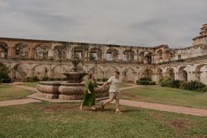 Couple dancing near a stone fountain in the courtyard of ancient ruins with arched walls and lush green lawn.