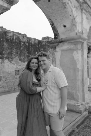 Black and white photo of a smiling couple embracing near stone arches at an ancient ruins landmark.