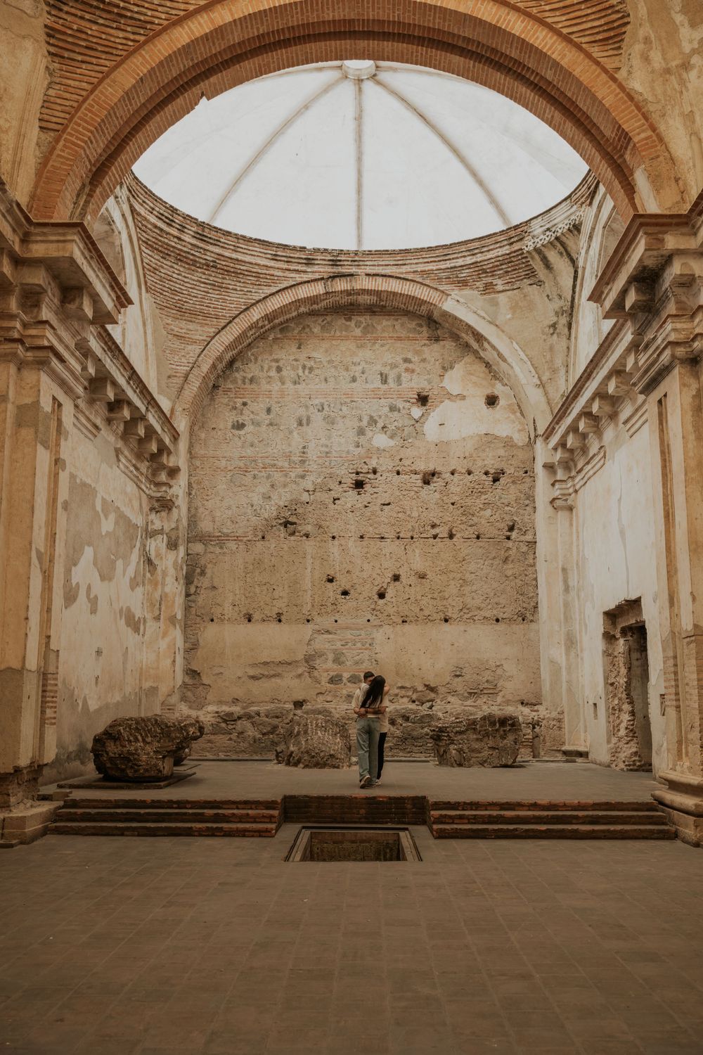 Person standing inside ancient ruined church with grand arch, dome, and weathered stone walls in Antigua, Guatemala.