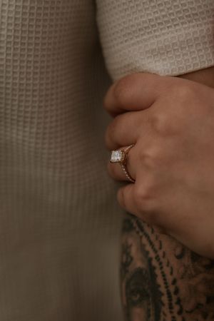 Close-up of a gold engagement ring with emerald-cut diamond on a hand with tattoos, against a textured cream background.